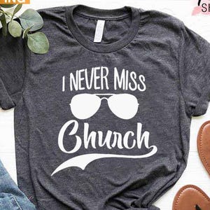 May include: Dark gray t-shirt with the text "I NEVER MISS Church" in white, along with sunglasses graphic. The shirt is paired with blue jeans and brown sandals. The words "NEXT DAY SHIPPING" are in a yellow box.