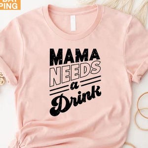 May include: A light pink t-shirt with the text "MAMA NEEDS a Drink" in black. The shirt has a crew neck and short sleeves. The words "NEXT DAY SHIPPING" are in a yellow box in the upper left corner.