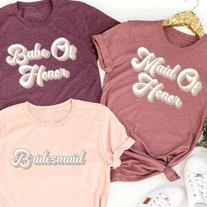 May include: Three short-sleeved t-shirts in heather maroon, heather mauve, and heather peach. The shirts feature the phrases "Babe Of Honor", "Maid Of Honor", and "Bridesmaid" in a retro font. White sneakers and a gold hanger are also visible.
