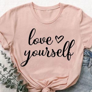 May include: A light pink t-shirt with the words "love yourself" in black script. The shirt is knotted at the waist and paired with denim shorts. The words "heather peach" are in a small circle in the upper left corner.