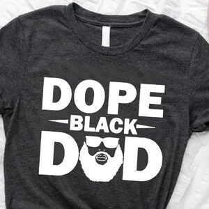 May include: A dark gray t-shirt with the text "Dope Black Dad" in white letters. The text is stylized with a beard and sunglasses.