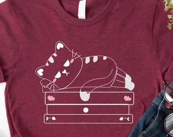 May include: A maroon t-shirt with a white outline of a cat sleeping on a stack of books. The text "CUSTOM TRENDY SHIRTS" is written in pink on a white background.