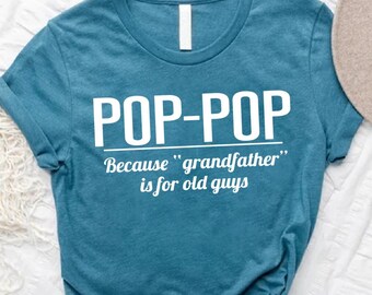May include: Teal t-shirt with the text "POP-POP" above the text "Because "grandfather" is for old guys" in white. A tan hat with a brown band and green leaves are in the background. The shirt is on a white surface.