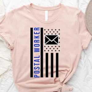 May include: A light pink t-shirt featuring a graphic design resembling a flag. The design includes the text "POSTAL WORKER" in blue, black stripes, stars, and an envelope icon. A hat and greenery are in the background.