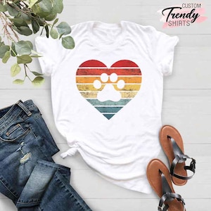May include: White t-shirt featuring a heart-shaped design with a paw print in the center. The heart has a retro color scheme of red, orange, yellow, and blue stripes. The text "Trendy Shirts" is printed in cursive.