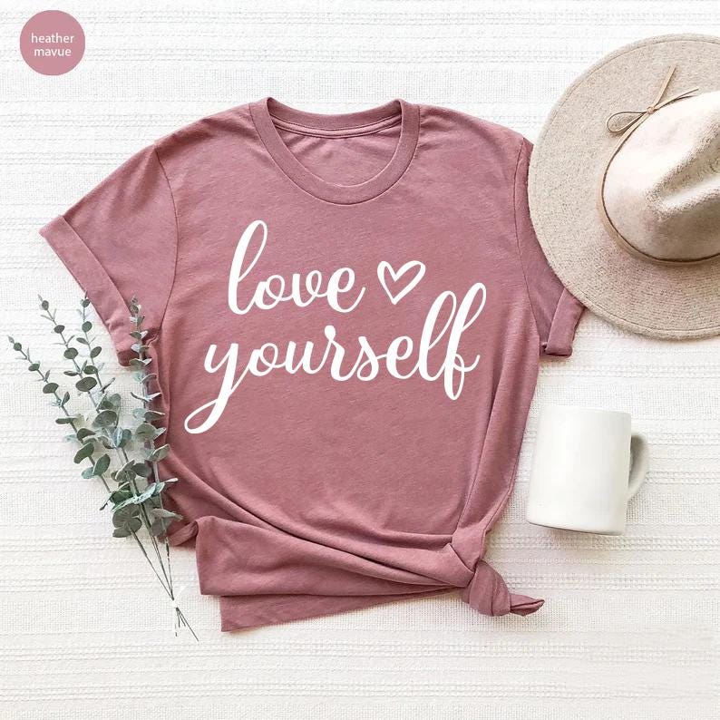 May include: A dusty rose-colored t-shirt with the words "love yourself" in white script, with a heart symbol. The shirt is styled with a knot at the bottom. A white mug and a beige hat are also in the image.