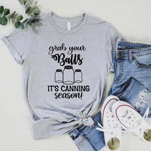 May include: A light gray t-shirt with the text "grab your Balls, it's canning season!" in black script. The shirt features a graphic of three canning jars. The shirt is paired with blue jeans and white sneakers.