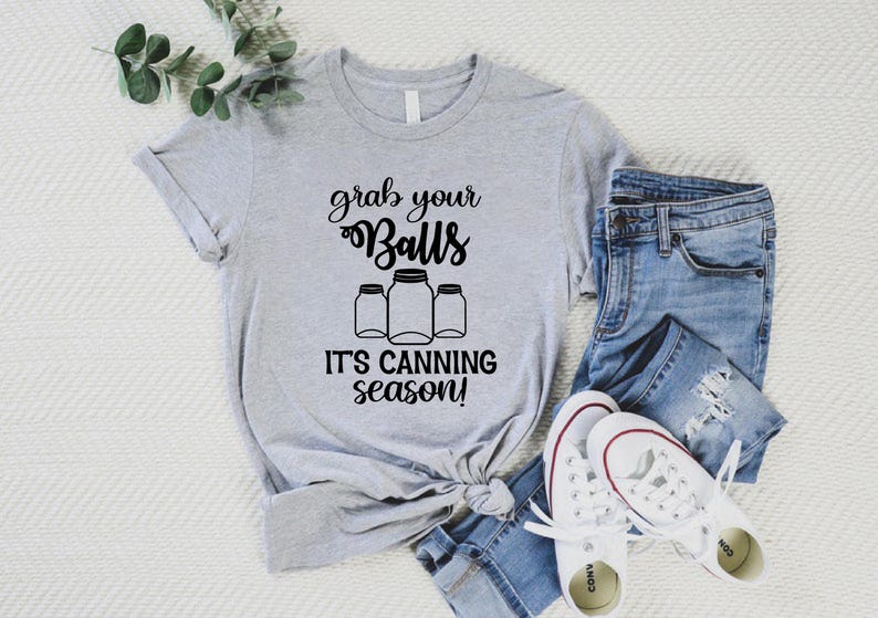 May include: A light gray t-shirt with the text "grab your Balls, it's canning season!" in black script. The shirt features a graphic of three canning jars. The shirt is paired with blue jeans and white sneakers.