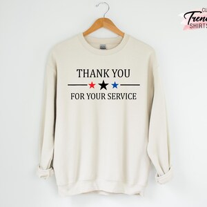 May include: Cream-colored sweatshirt with the text "THANK YOU FOR YOUR SERVICE" in black, with a black line and red, black, and blue stars. The sweatshirt is hanging on a wooden hanger.