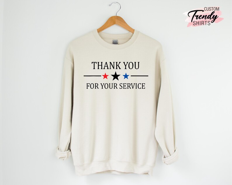 May include: Cream-colored sweatshirt with the text "THANK YOU FOR YOUR SERVICE" in black, with a black line and red, black, and blue stars. The sweatshirt is hanging on a wooden hanger.