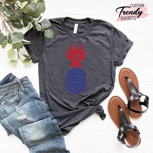 May include: A dark gray t-shirt with a red and blue pineapple design. The shirt is made of a soft, comfortable material and is perfect for everyday wear.