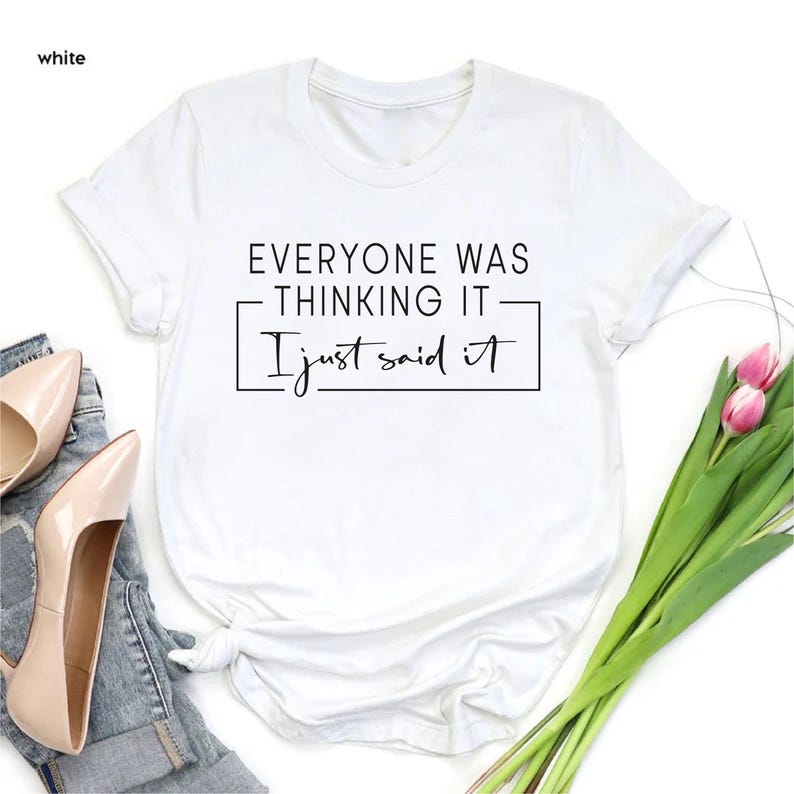 May include: White t-shirt with the text "EVERYONE WAS THINKING IT - I just said it" in black. The shirt is styled with a pair of beige heels, blue jeans, and a bouquet of pink tulips.