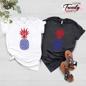 May include: Two t-shirts with a pineapple design. The pineapple is red, white, and blue. One shirt is white and the other is black. CUSTOM Trendy SHIRTS is written in pink at the top of the image.