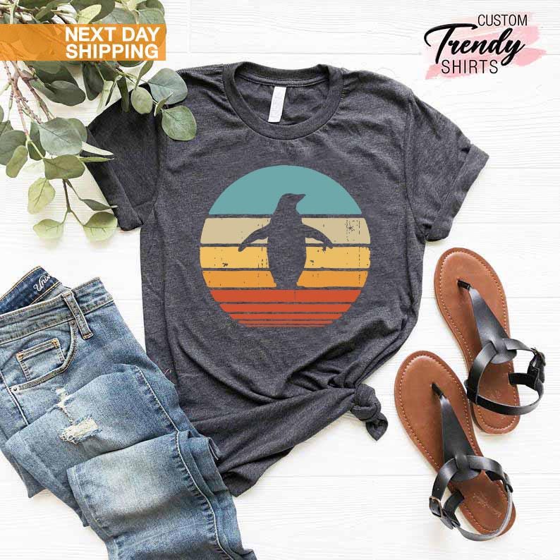 May include: A dark gray t-shirt with a retro-style sunset graphic featuring a penguin silhouette. The graphic has a blue, turquoise, yellow, orange, and red color scheme. The t-shirt is displayed on a white surface with a pair of brown sandals and a pair of blue jeans.