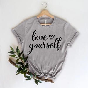 May include: A gray t-shirt with the words "love yourself" in black script, with a heart symbol. The shirt is hanging on a wooden hanger. A sprig of greenery is placed on the lower left side of the shirt.