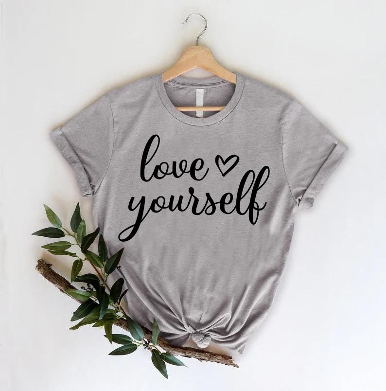 May include: A gray t-shirt with the words "love yourself" in black script, with a heart symbol. The shirt is hanging on a wooden hanger. A sprig of greenery is placed on the lower left side of the shirt.