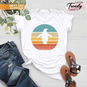 May include: White t-shirt with a retro-style graphic of a penguin standing in front of a sunset. The graphic has a distressed look with a teal, beige, yellow, orange, and red color scheme.