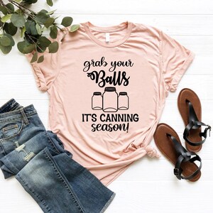 May include: A light pink t-shirt with the text "grab your Balls" above three jar illustrations and the phrase "IT'S CANNING season!". The shirt is paired with blue jeans and black sandals.