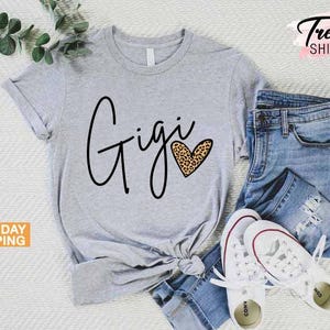 May include: Gray t-shirt with the word "Gigi" in black script and a leopard print heart. The shirt is shown on a model wearing blue jeans and white sneakers.