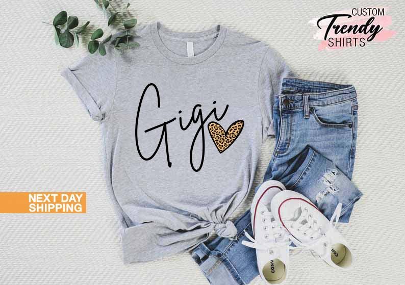 May include: Gray t-shirt with the word "Gigi" in black script and a leopard print heart. The shirt is shown on a model wearing blue jeans and white sneakers.