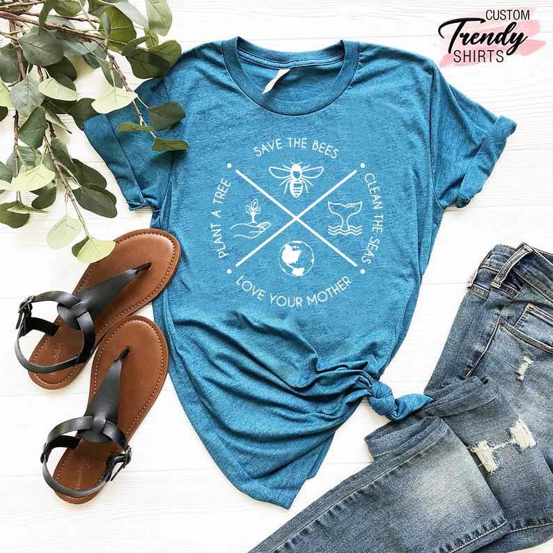 Op de afbeelding: Een tealblauw t-shirt met een wit grafisch ontwerp met een cirkel met de tekst "Save the Bees", "Plant a Tree", "Love Your Mother" en "Clean the Seas" met een bij, een boom, een walvis en een wereldbol in de cirkel.