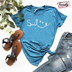 Op de afbeelding: Een tealblauw t-shirt met het woord "Smile" geschreven in wit cursief met een smiley ernaast getekend. Het shirt ligt op een wit oppervlak met een paar bruine sandalen en een blauwe jeans.