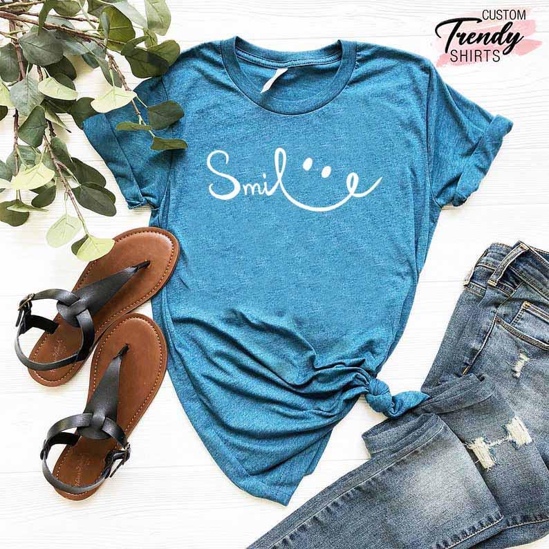 Op de afbeelding: Een tealblauw t-shirt met het woord "Smile" geschreven in wit cursief met een smiley ernaast getekend. Het shirt ligt op een wit oppervlak met een paar bruine sandalen en een blauwe jeans.