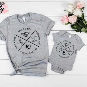 Op de afbeelding: Een set van twee grijze T-shirts met een zwart-wit grafisch ontwerp. Het grotere shirt heeft de tekst "Save the Bees, Plant a Tree, Clean the Seas, Love Your Mother". Het kleinere shirt heeft hetzelfde ontwerp, maar is kleiner.