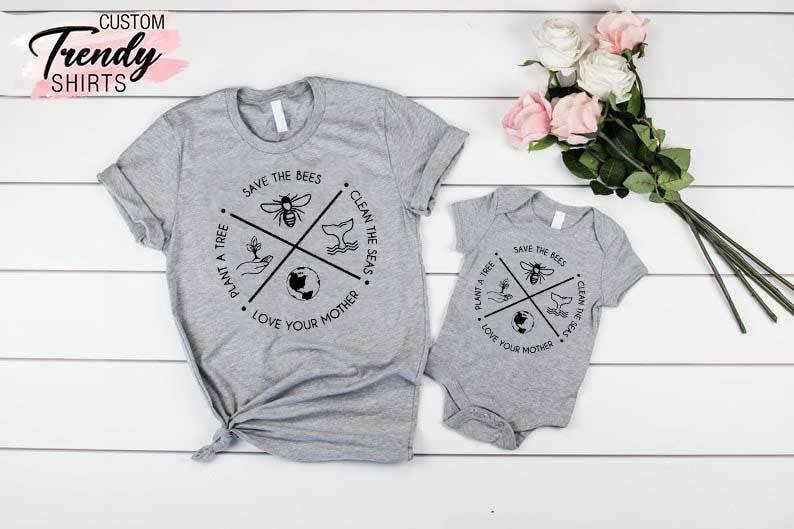Op de afbeelding: Een set van twee grijze T-shirts met een zwart-wit grafisch ontwerp. Het grotere shirt heeft de tekst "Save the Bees, Plant a Tree, Clean the Seas, Love Your Mother". Het kleinere shirt heeft hetzelfde ontwerp, maar is kleiner.