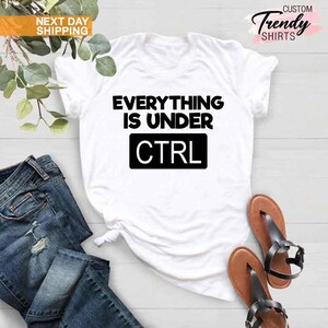 K&ouml;nnte beinhalten: Wei&szlig;es T-Shirt mit dem schwarzen Text "EVERYTHING IS UNDER CTRL". Das Shirt wird mit blauen Jeans und braunen Sandalen kombiniert. Das Bild enth&auml;lt auch einen Zweig mit Gr&uuml;n.