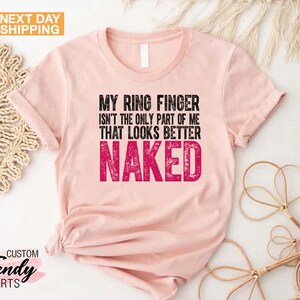 May include: A light pink t-shirt with the text "MY RING FINGER ISN'T THE ONLY PART OF ME THAT LOOKS BETTER NAKED" in black and pink. The shirt has a crew neck and short sleeves. The words "NEXT DAY SHIPPING" are in the top left corner.