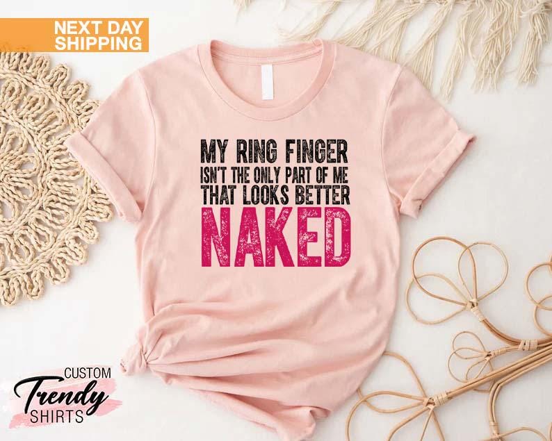 May include: A light pink t-shirt with the text "MY RING FINGER ISN'T THE ONLY PART OF ME THAT LOOKS BETTER NAKED" in black and pink. The shirt has a crew neck and short sleeves. The words "NEXT DAY SHIPPING" are in the top left corner.