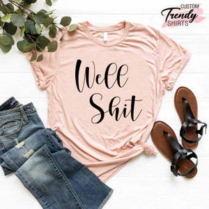 May include: A light pink t-shirt with the text "Well Shit" printed in black cursive font.