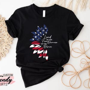 May include: Black t-shirt with a red, white, and blue American flag design in the shape of a flower. The text "Land of the Free Because of the Brave" is printed on the shirt.