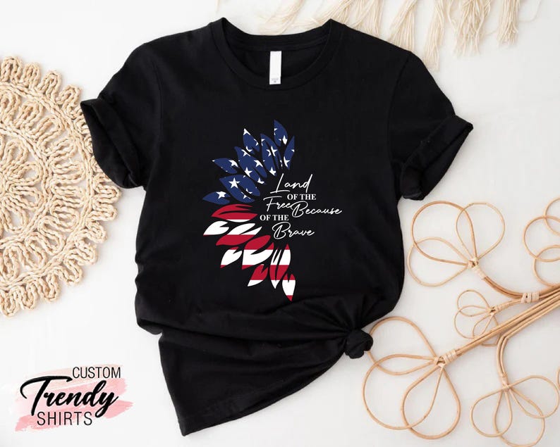 May include: Black t-shirt with a red, white, and blue American flag design in the shape of a flower. The text "Land of the Free Because of the Brave" is printed on the shirt.