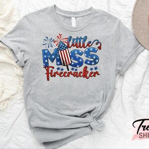 May include: Gray t-shirt with a red, white, and blue firework graphic and the text "little miss firecracker".