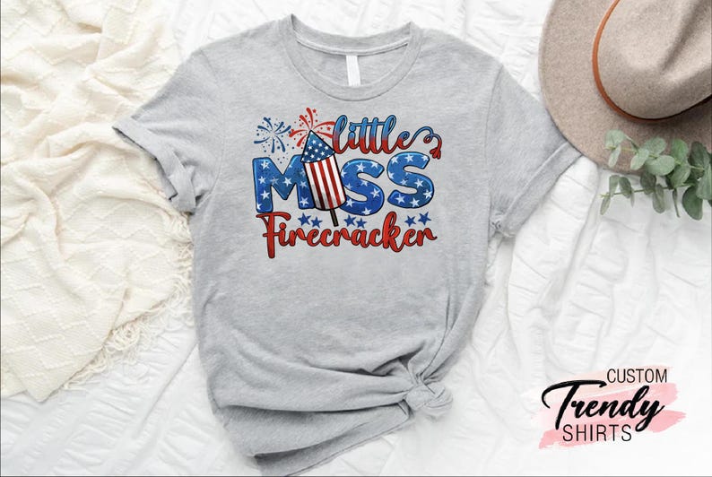 May include: Gray t-shirt with a red, white, and blue firework graphic and the text "little miss firecracker".