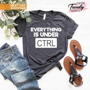 K&ouml;nnte beinhalten: Dunkelgraues T-Shirt mit dem wei&szlig;en Text "EVERYTHING IS UNDER CTRL". Das Shirt wird mit blauen Jeans und braunen Sandalen kombiniert. Auch ein Zweig mit Gr&uuml;n ist im Bild zu sehen.