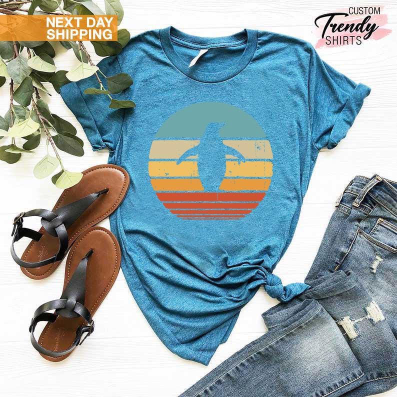 May include: A teal blue t-shirt with a retro-style graphic of a penguin silhouette in front of a sunset. The graphic is in shades of orange, yellow, and red.