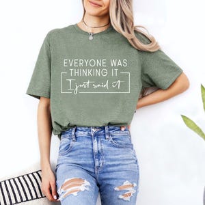 May include: An olive green t-shirt with the text "EVERYONE WAS THINKING IT I just said it" in white. The shirt is paired with blue jeans. The image is of a person wearing the shirt.