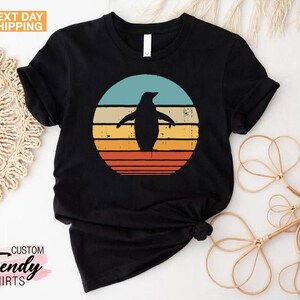 May include: Black t-shirt with a retro-style graphic of a penguin standing in front of a sunset. The graphic has a red, orange, yellow, and blue color scheme.