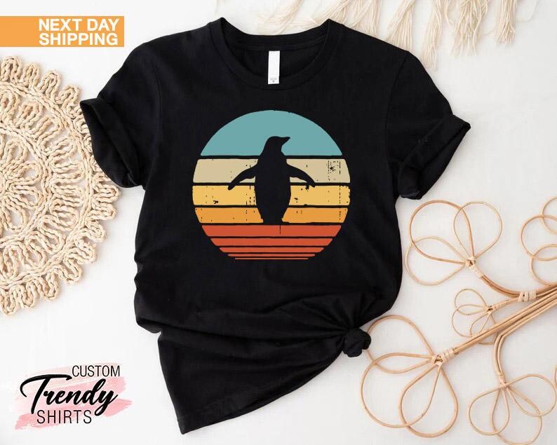 May include: Black t-shirt with a retro-style graphic of a penguin standing in front of a sunset. The graphic has a red, orange, yellow, and blue color scheme.