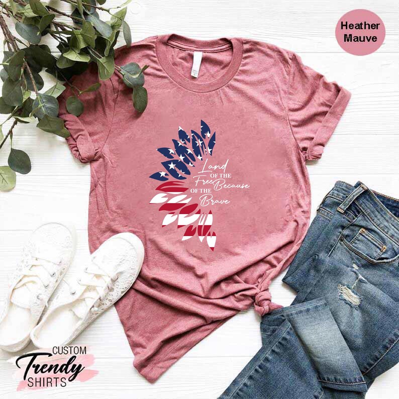 May include: A heather mauve t-shirt with a graphic of a stylized American flag in the shape of a sunflower. The text "Land of the Free Because of the Brave" is printed on the shirt.