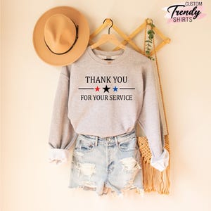 May include: A light gray sweatshirt with the text "THANK YOU FOR YOUR SERVICE" in black, red, and blue. The sweatshirt is paired with denim shorts and a tan hat.