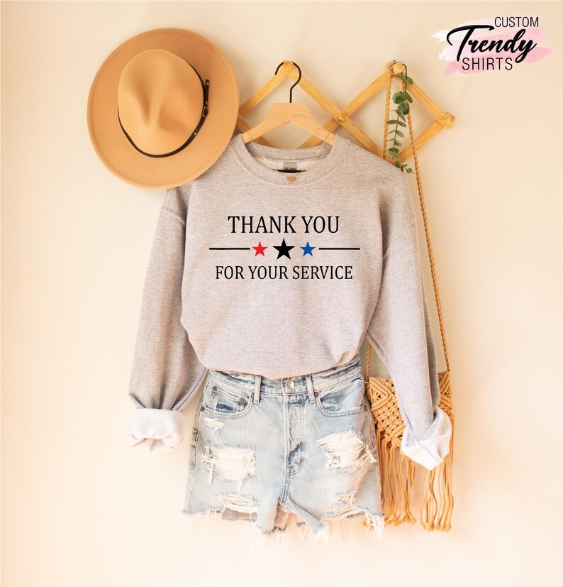May include: A light gray sweatshirt with the text "THANK YOU FOR YOUR SERVICE" in black, red, and blue. The sweatshirt is paired with denim shorts and a tan hat.