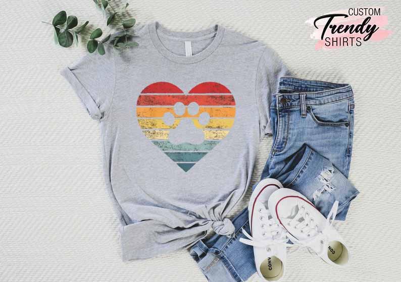 Op de afbeelding: Een grijze t-shirt met een hartvormig ontwerp met een pootafdruk in het midden. Het hart heeft een retro kleurenschema van rode, oranje, gele en blauwe strepen. De tekst "CUSTOM Trendy SHIRTS" staat in de rechterbovenhoek.