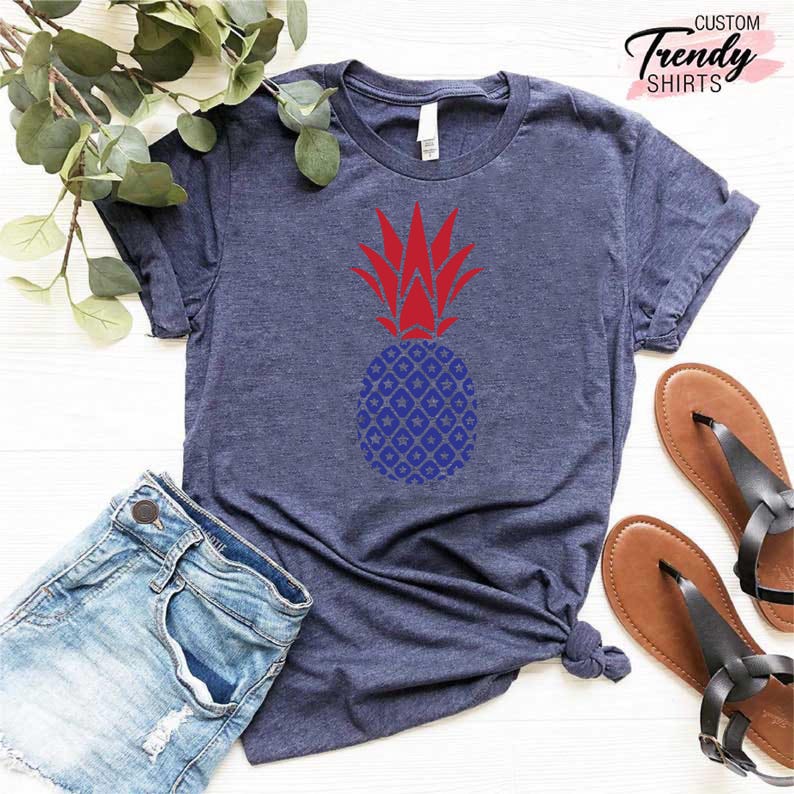 May include: A navy blue t-shirt with a red and blue pineapple design. The pineapple has a red top and a blue patterned body.