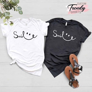 Op de afbeelding: Twee T-shirts met het woord "Smile" en een smiley-ontwerp. Een shirt is wit en het andere is zwart.