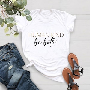 May include: White t-shirt with the text "HUMAN KIND be both" in black lettering.
