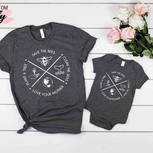 Op de afbeelding: Een set van twee grijze t-shirts met een wit grafisch ontwerp. Het grotere shirt heeft een rond ontwerp met de tekst "Save the Bees", "Plant a Tree", "Clean the Seas" en "Love Your Mother". Het kleinere shirt is een baby-romper met hetzelfde ontwerp.